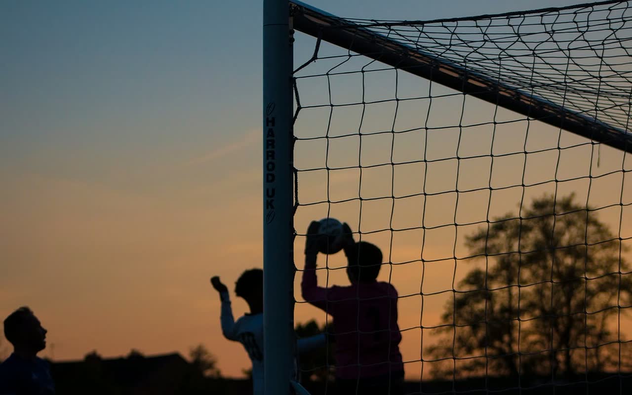 Crianças em silhueta junto à trave de um campo de futebol ao pôr do sol